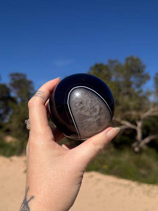 Hand holding a polished black and grey banded crystal sphere against a bright blue sky with sandy ground and green trees in the background.