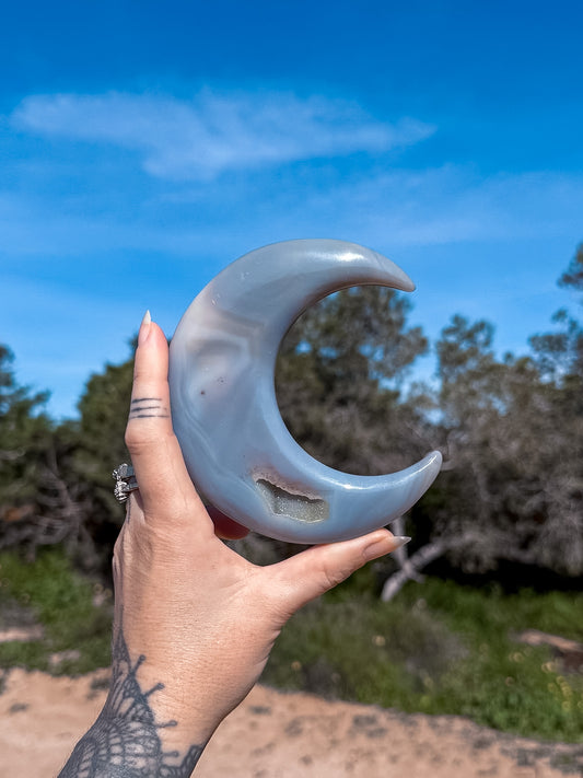 Hand holding a large, polished blue agate crystal crescent moon against a natural background with trees and blue sky.