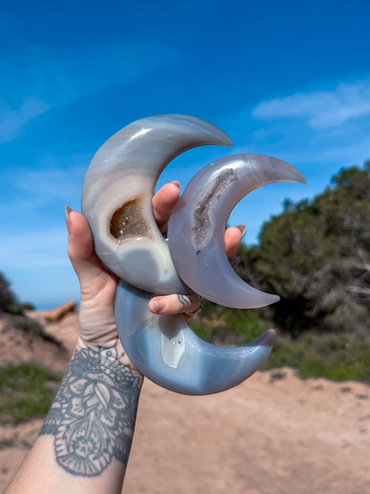 Hand holding three large, curved ble agate crystal moons against a blue sky and natural background