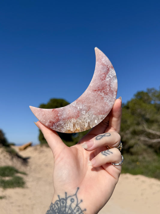 Hand holding a polished crescent pink amethyst moon carving made of pink and clear crystal with red inclusions against a bright blue sky and sandy coastal background.