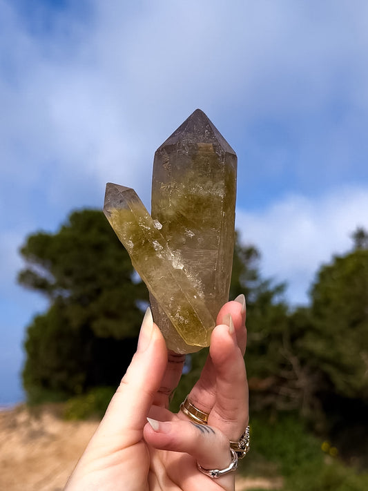 Hand holding a raw smoky citrine double point crystal against a natural background with trees and blue sky.