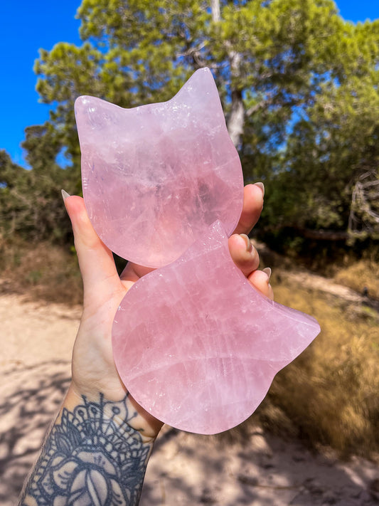 A hand holding two cat-shaped crsytal bowls made from rose quartz, with a background of a natural outdoor setting.