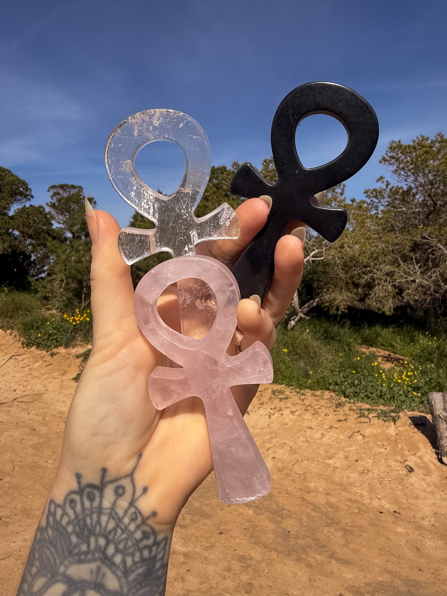 Hand holding three crystal carving of ankh's in rose quartz, clear quartz and obsidian against a natural background 