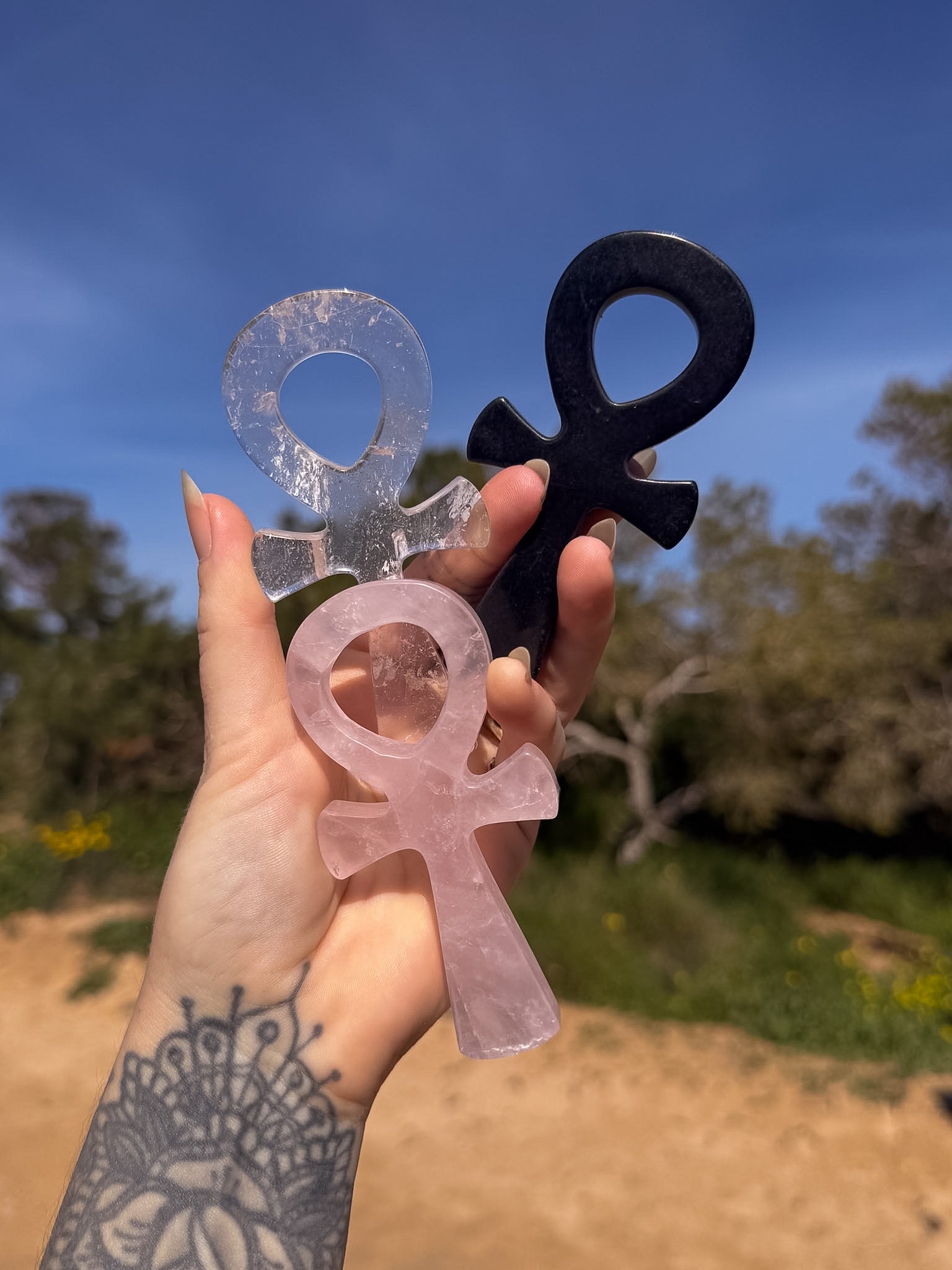 Hand holding three crystal carving of ankh's in rose quartz, clear quartz and obsidian against a natural background 