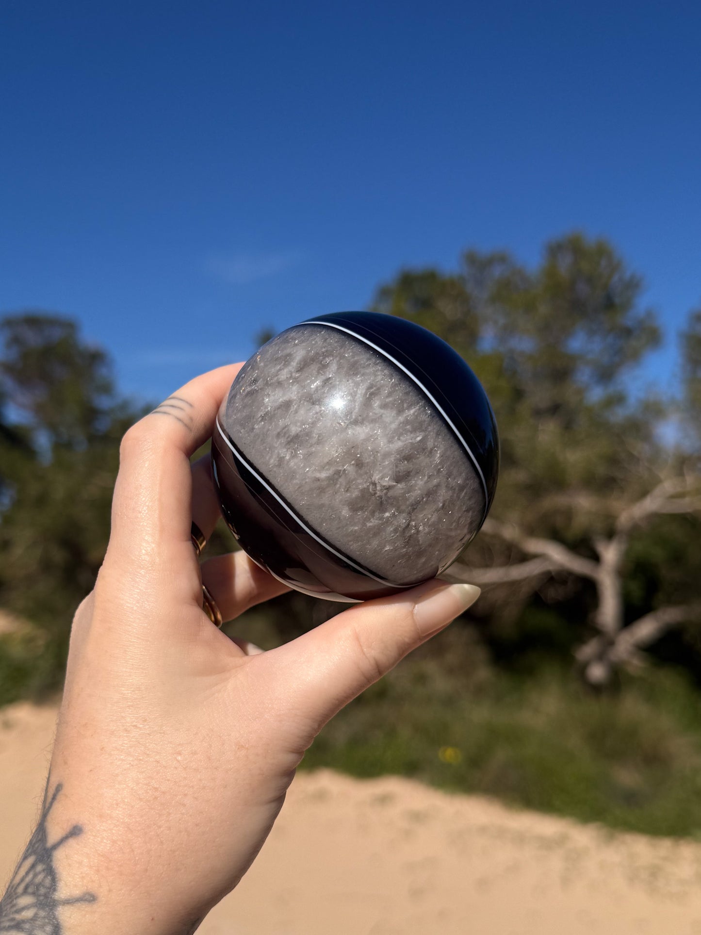 Hand holding a polished black and grey banded crystal sphere against a bright blue sky with sandy ground and green trees in the background.