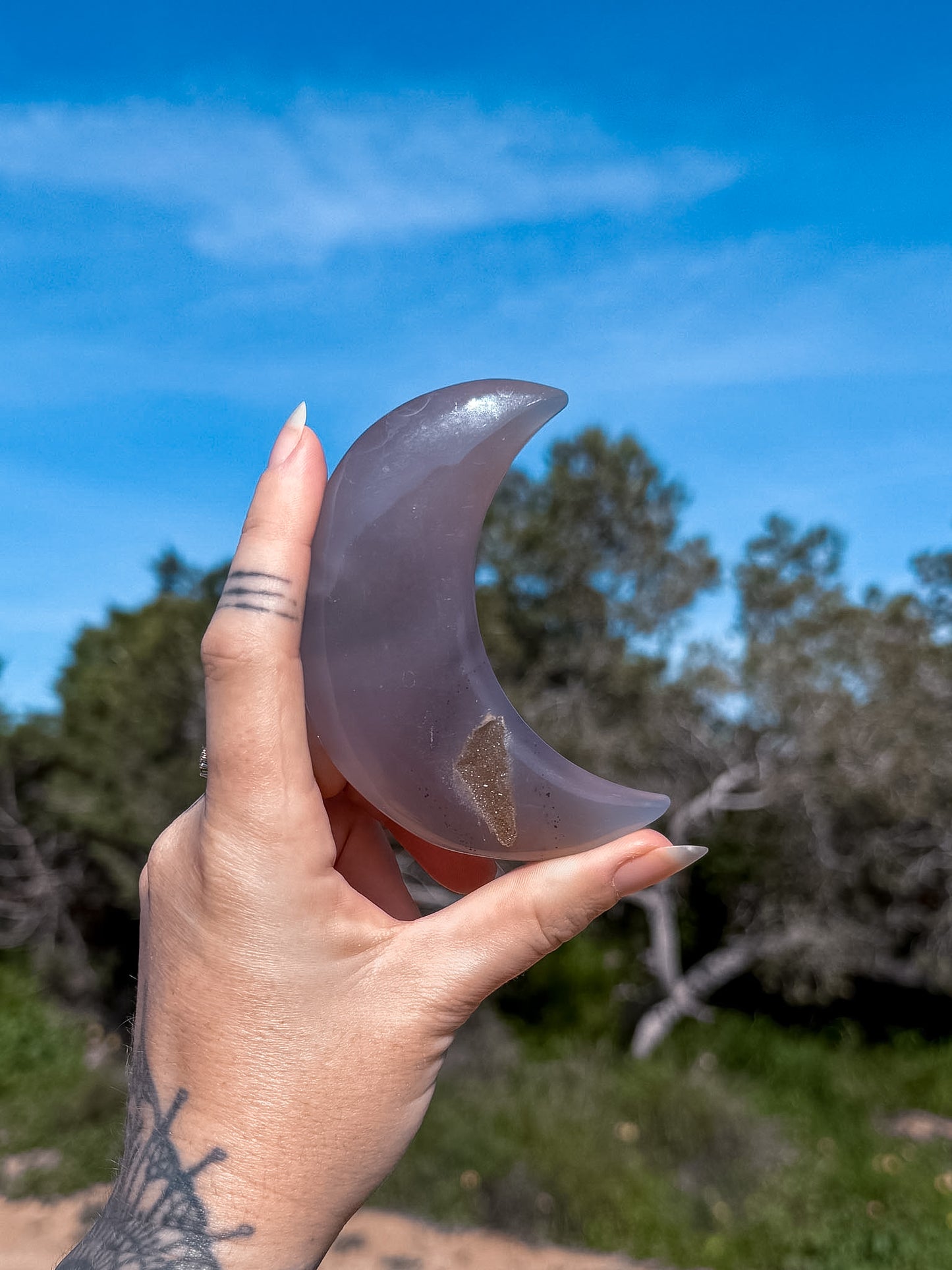 Hand holding a carved greyish blue agate crystal crescent moon against a natural background with trees and blue sky.