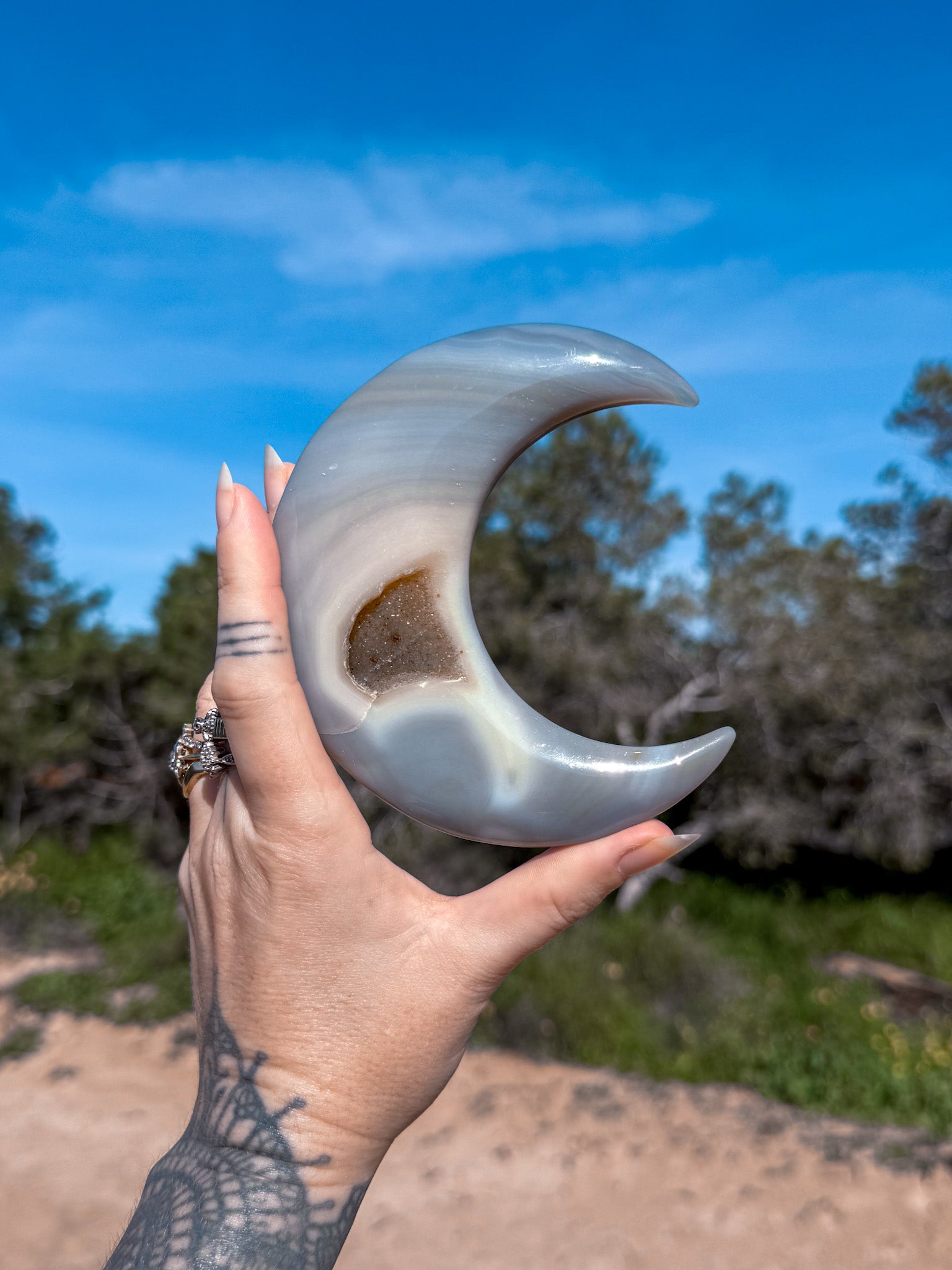 Hand holding a large, blue agate curved crescent moon stone against a natural background with trees and blue sky.