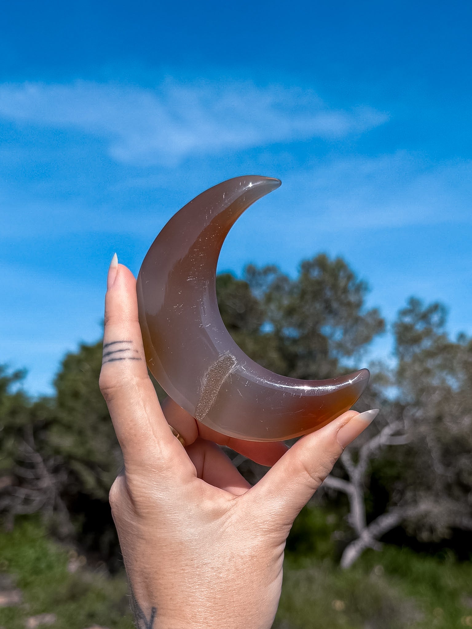 Hand holding a blue and brown agate crescent moon shaped stone against a blue sky with trees in the background