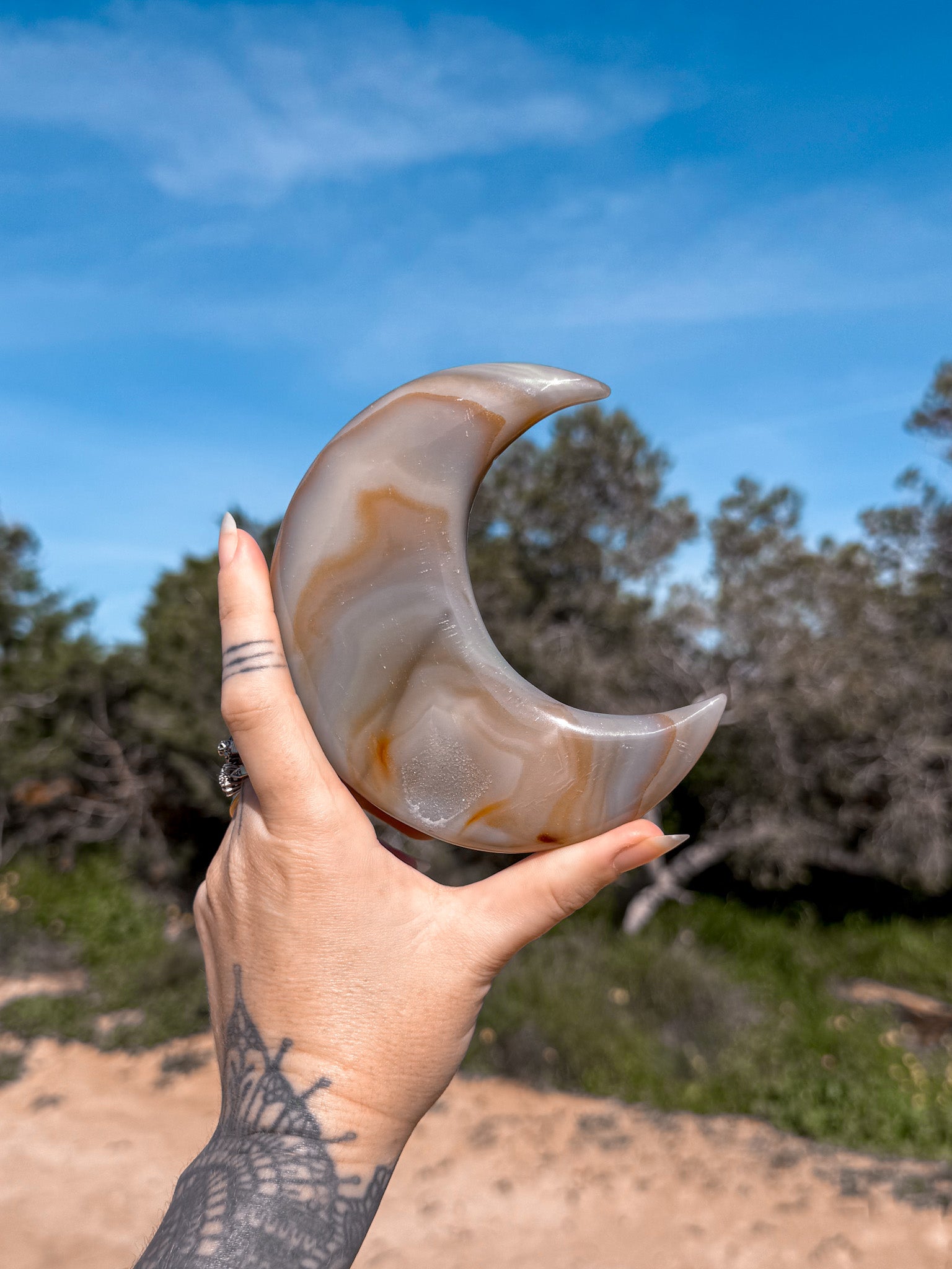 Hand holding a large, polished blue and golden agate crystal crescent moon against a natural background with trees and blue sky.