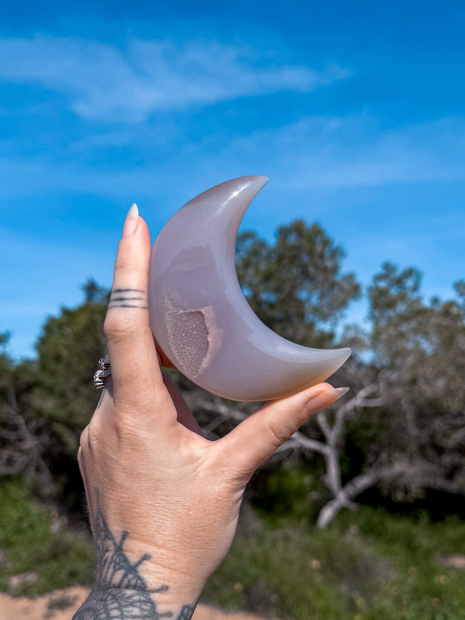 Hand holding a blue agate crystal  crescent moon-shaped stone against a blue sky with trees in the background