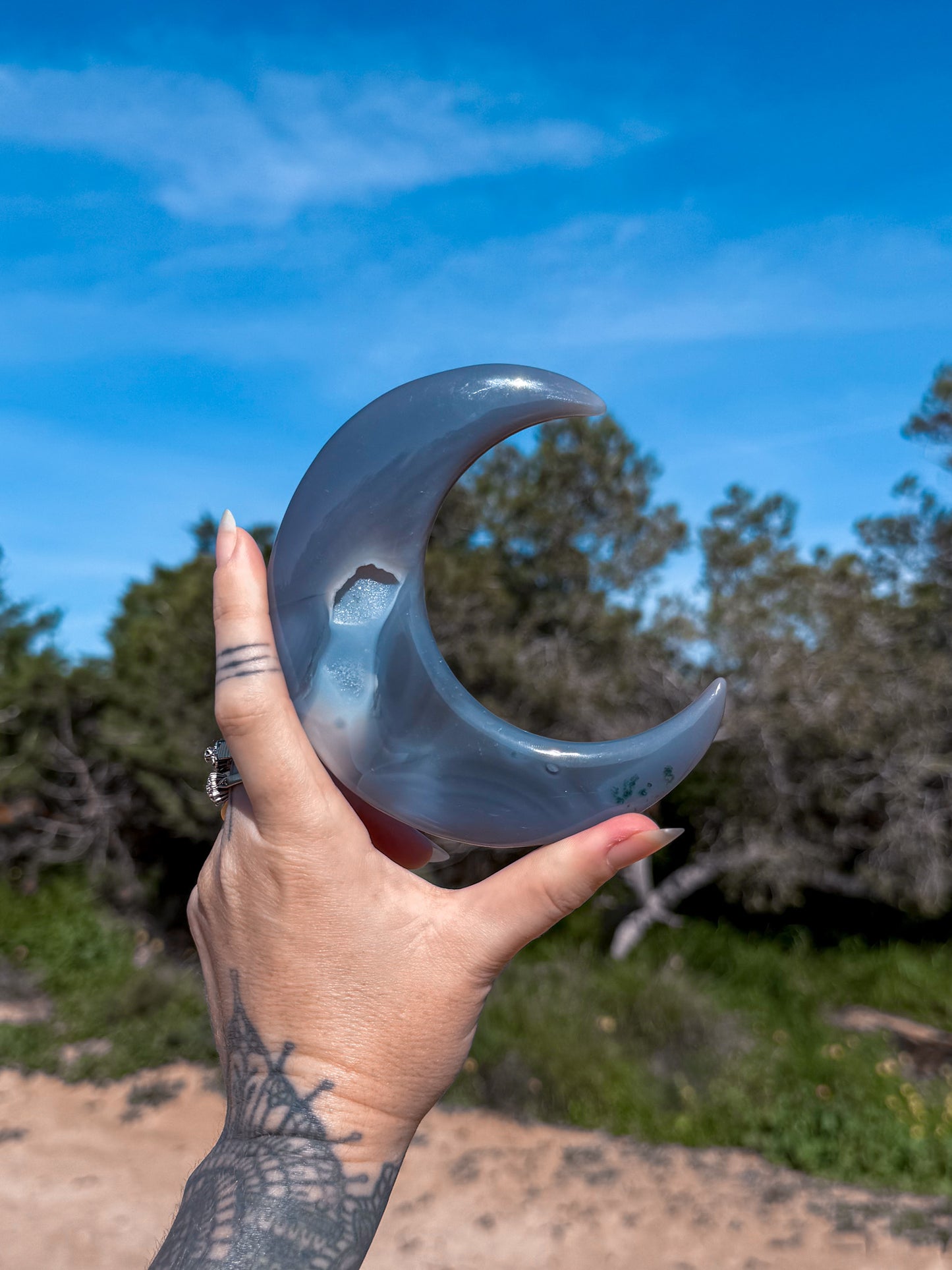 Hand holding a blue agate stone crescent moon against a natural background with trees and blue sky.