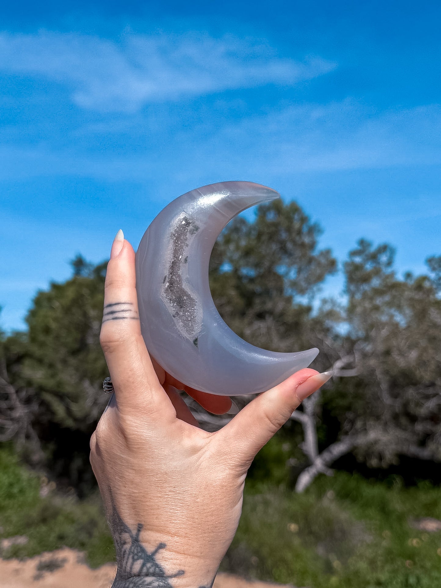 Hand holding a blue agate crescent-shaped stone with natural druzy against a natural background with trees and blue sky.