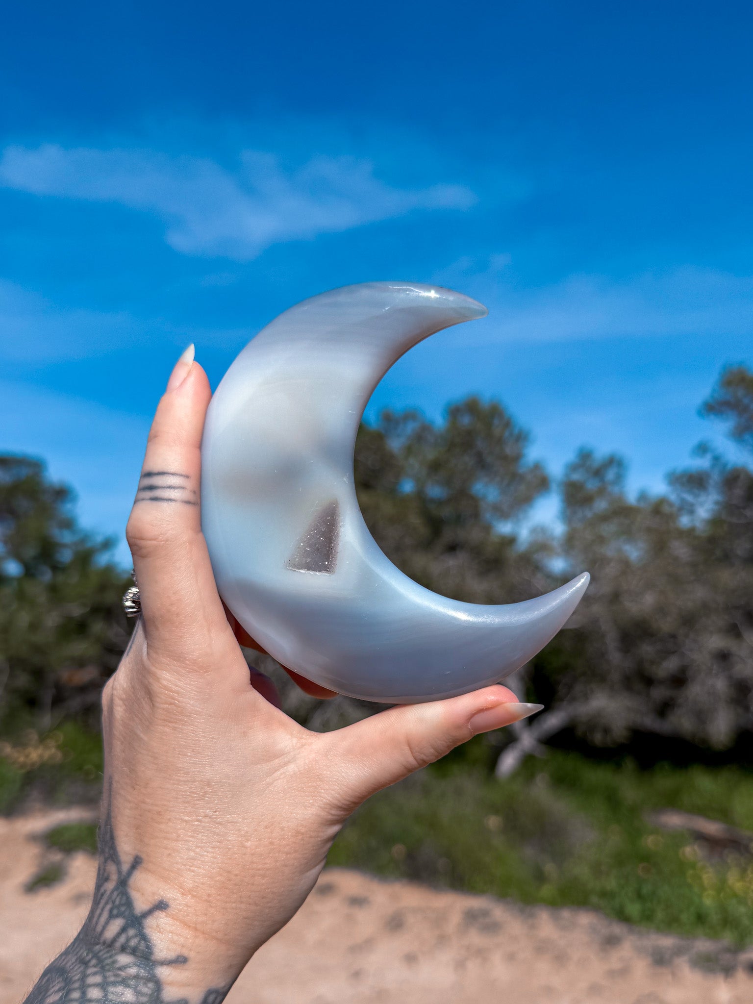 Hand holding a crescent moon-shaped blue agate crystal against a natural background with trees and blue sky.