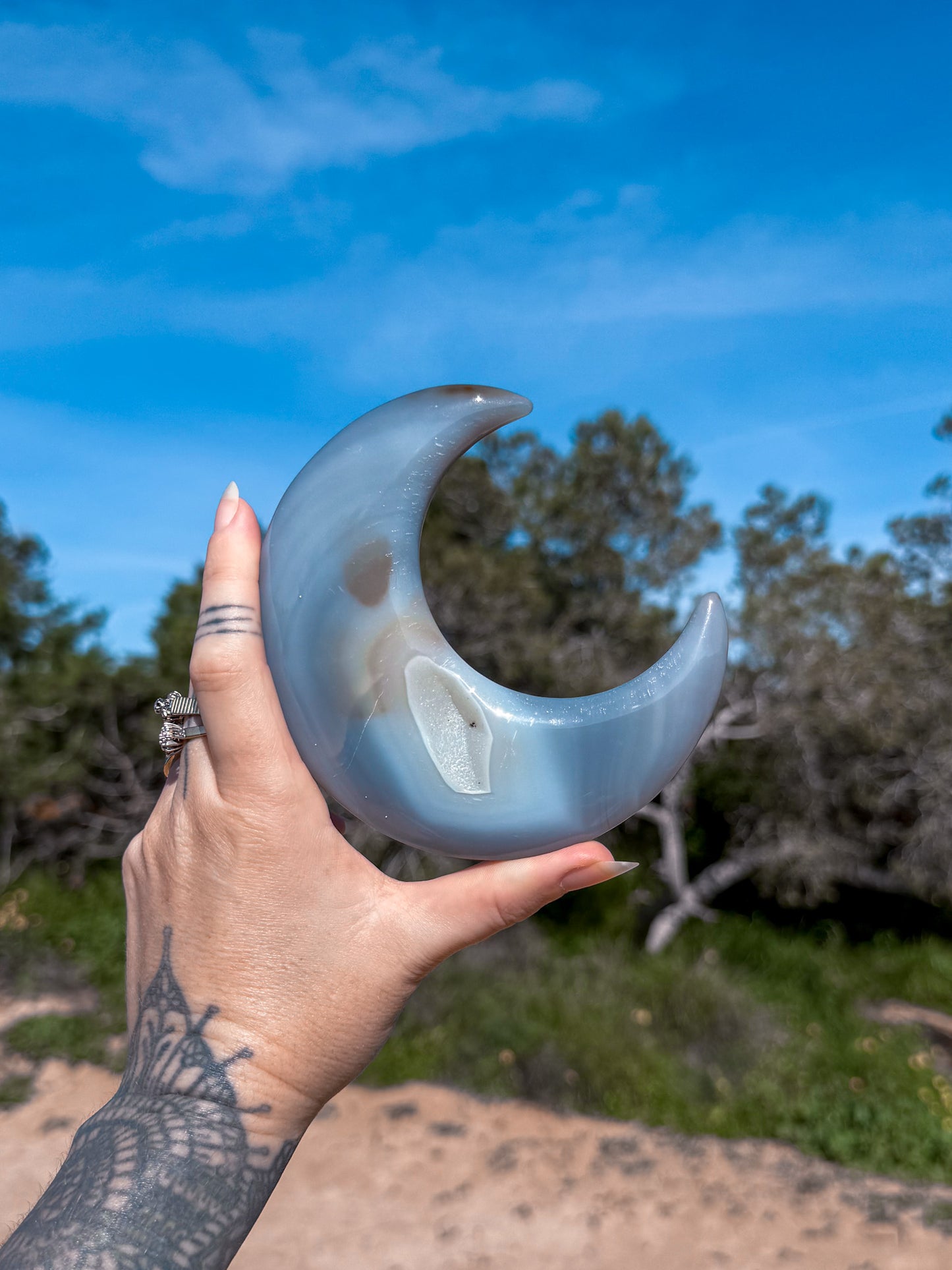 Hand holding a blue agate crescent moon crystal against a natural background with trees and sky.