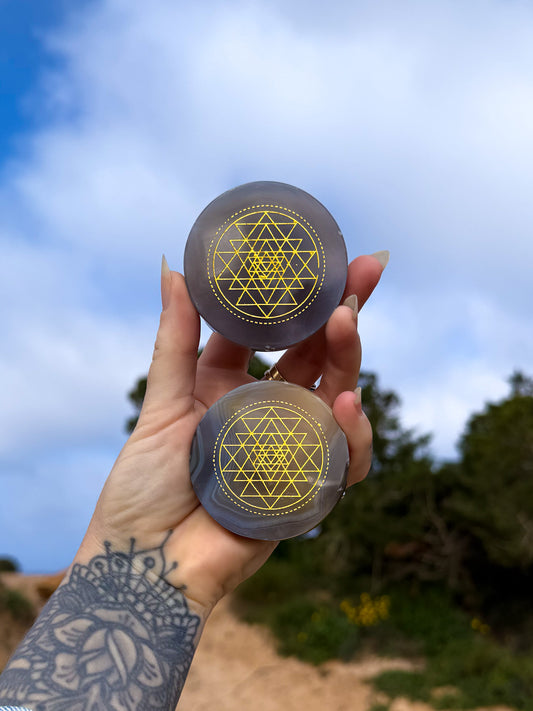 hand holding two blue agate crsytal discs with gold sri yantra geometric pattern printed on top against a blue sky and greenery background.