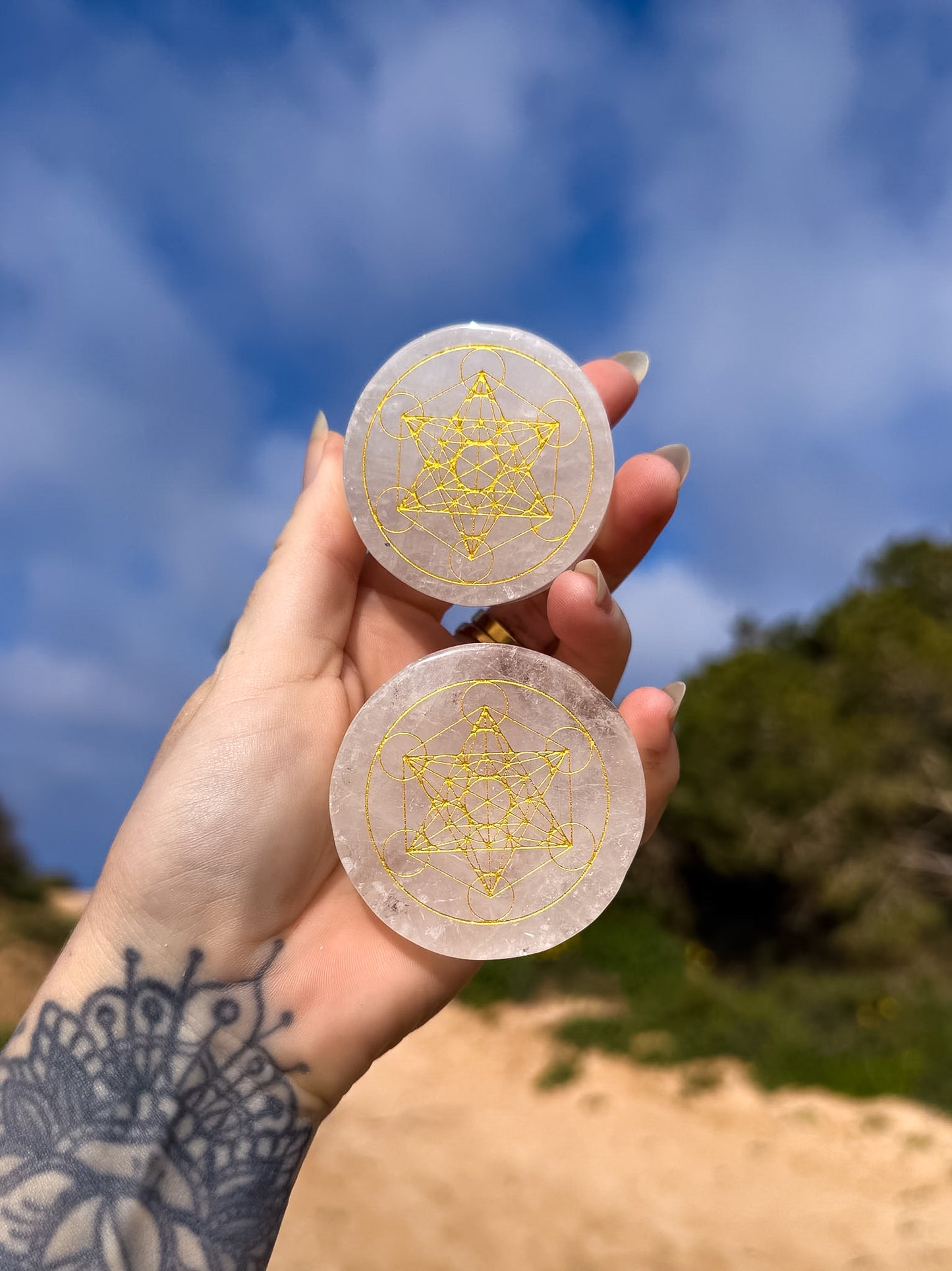 Hand holding two circular clear quartz with sacred geometric gold design, metatrons cube against a blue sky and green trees background