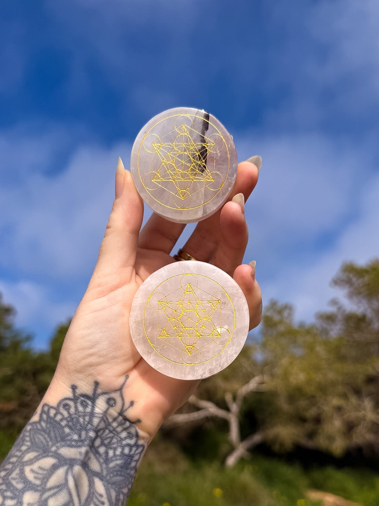 Hand holding two circular clear quartz with sacred geometric gold design, metatrons cube against a blue sky and green trees background