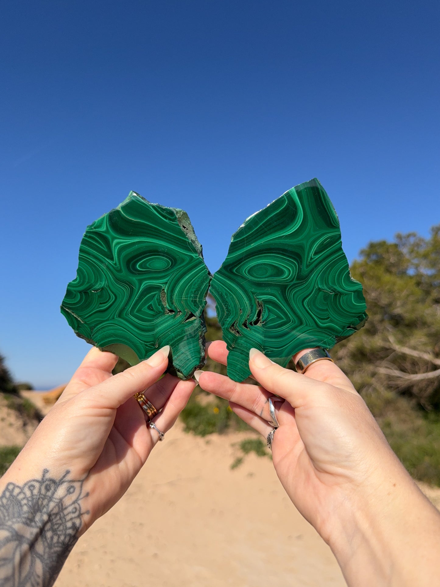 Two hands holding matching polished malachite slices with vivid green concentric banding against a bright blue sky and sandy coastal landscape background.