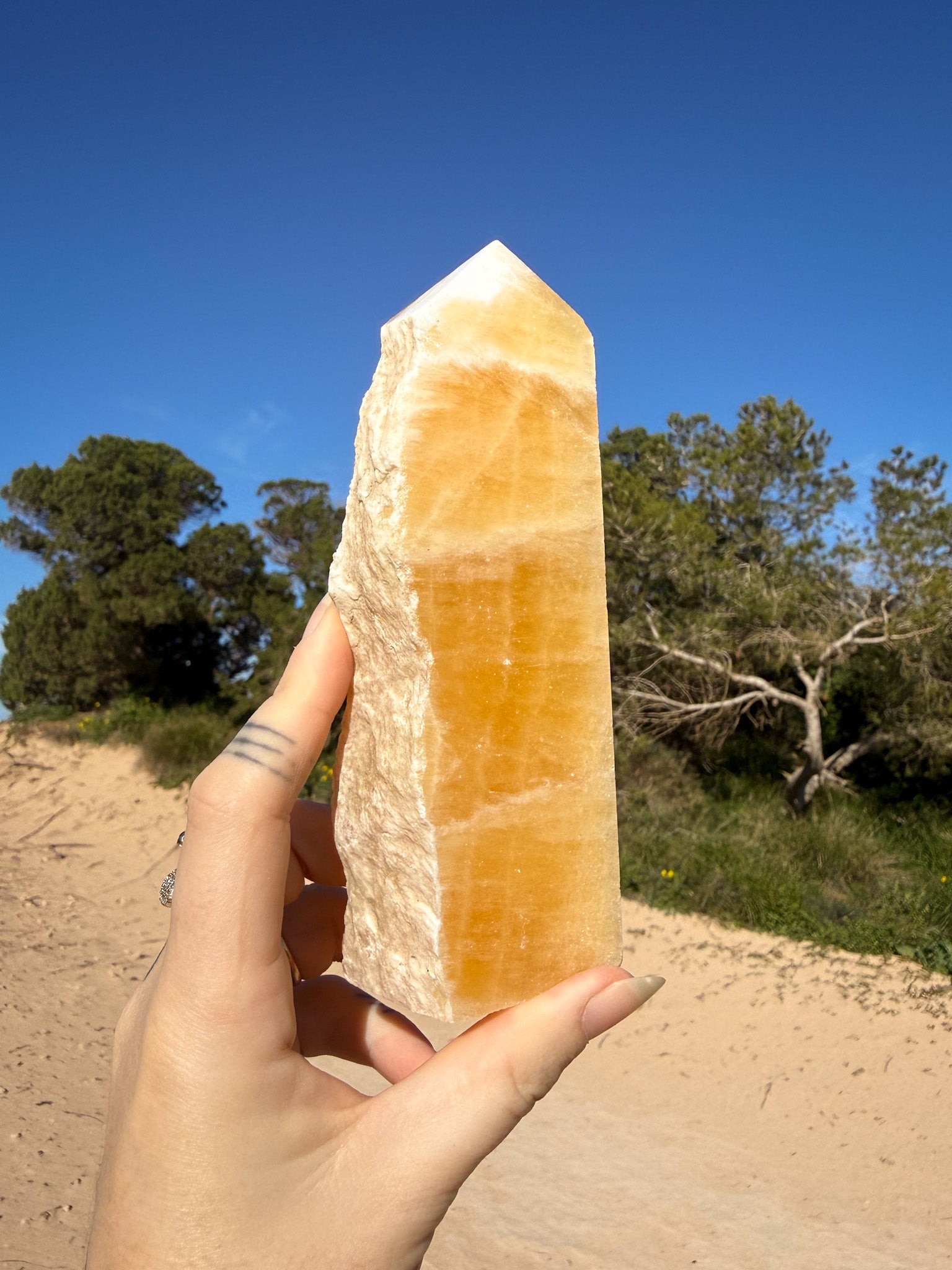 Hand holding a polished orange calcite tower with natural white banding against a bright blue sky and sandy coastal landscape background.