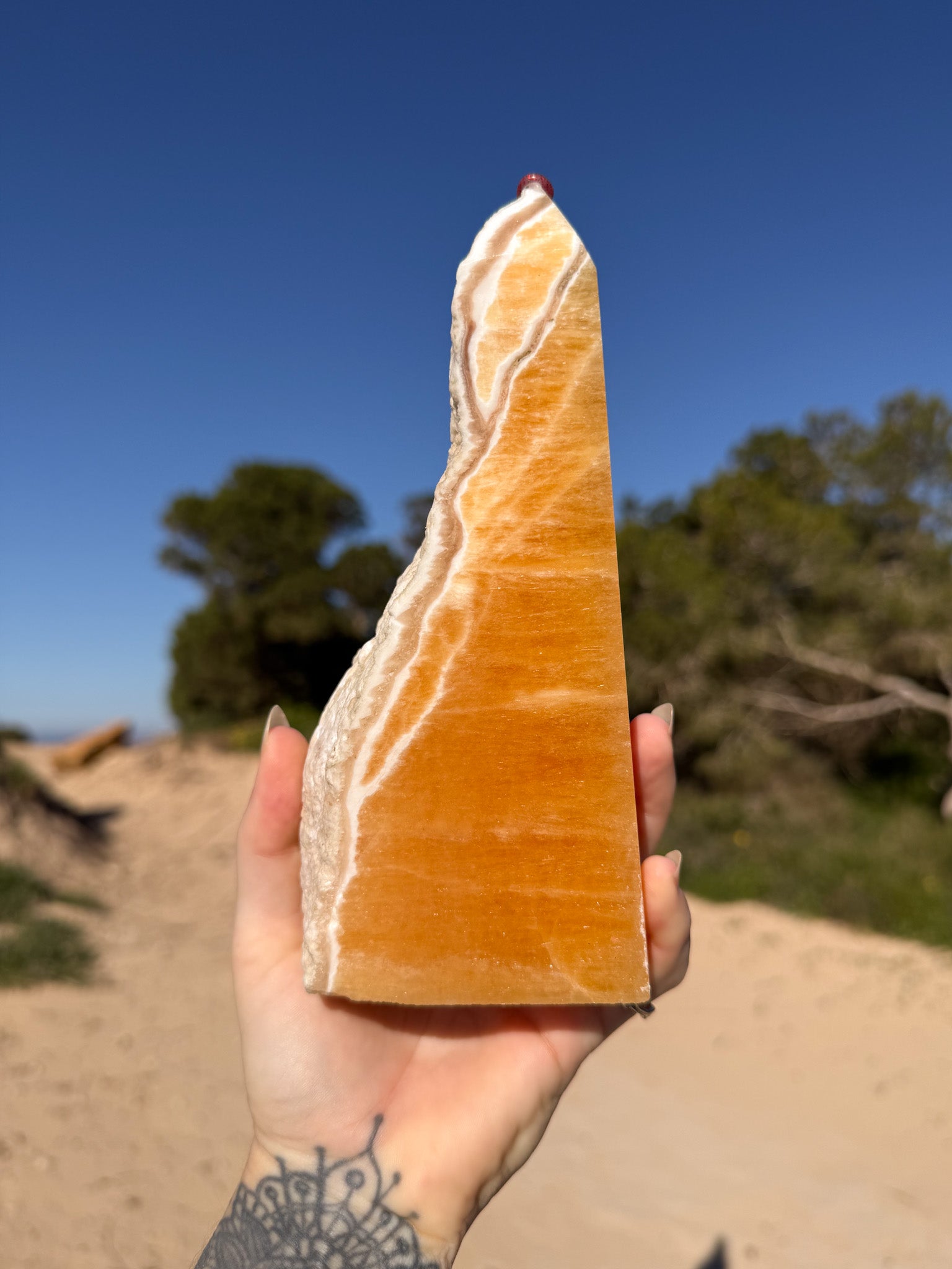 Hand holding a polished orange calcite tower with natural white banding against a bright blue sky and sandy coastal landscape background.