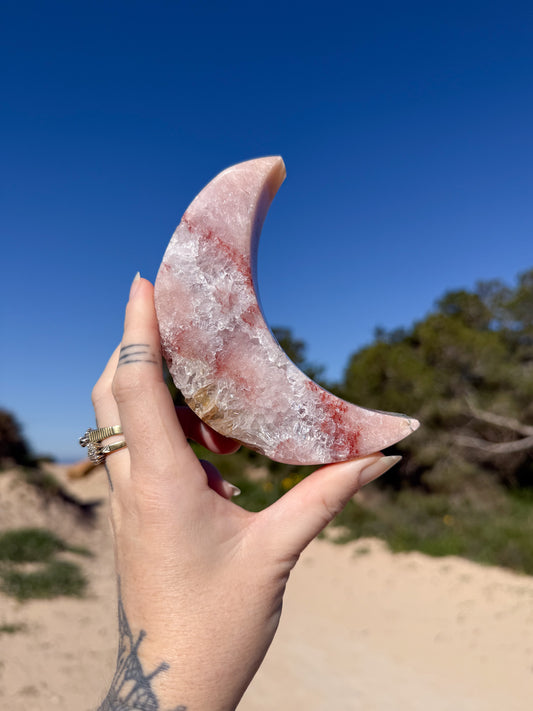 Hand holding a polished crescent pink amethyst moon carving made of pink and clear crystal with red inclusions against a bright blue sky and sandy coastal background.