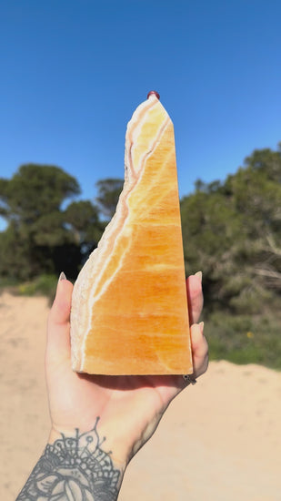 Hand holding a polished orange calcite tower with natural white banding against a bright blue sky and sandy coastal landscape background.