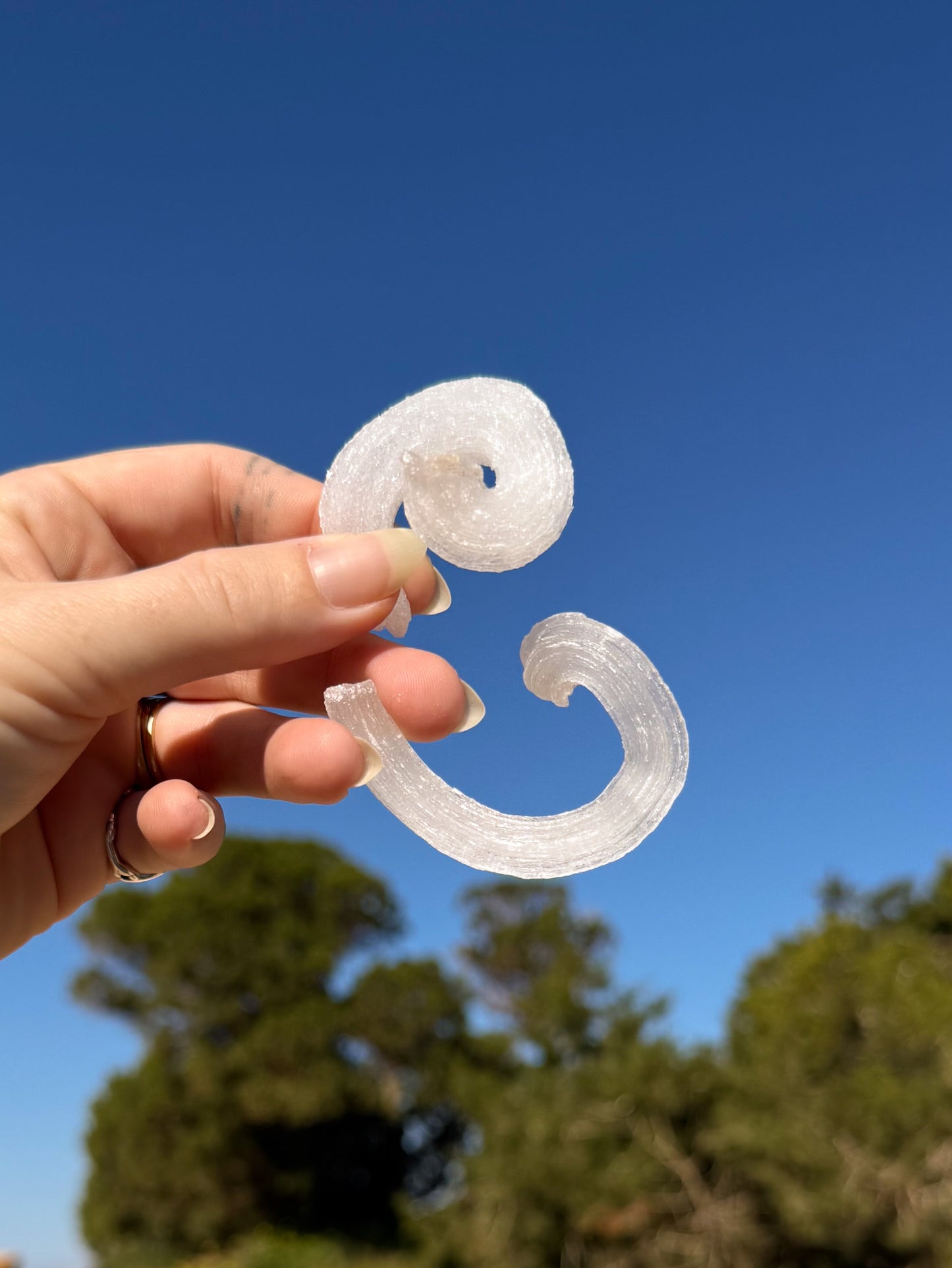 Two hands holding matching white sparkly raw rams horm selenite crystal against a bright blue sky with sandy ground and green trees in the background.