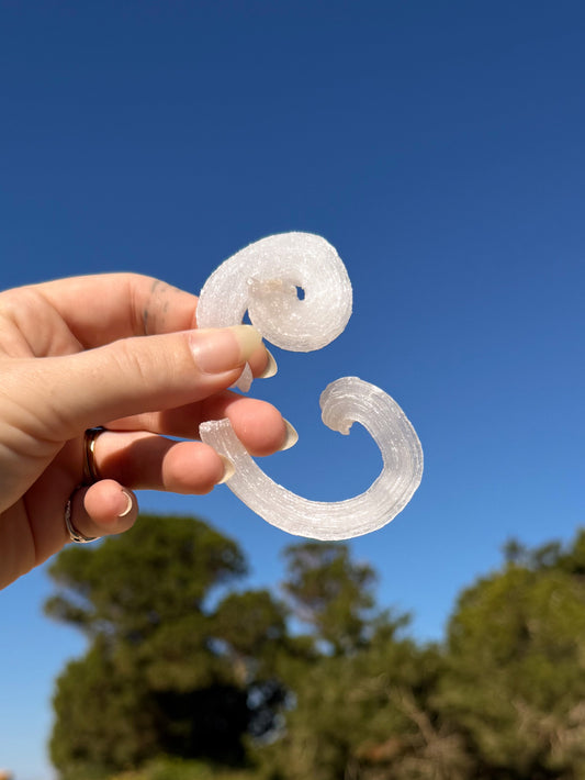 Two hands holding matching white sparkly raw rams horm selenite crystal against a bright blue sky with sandy ground and green trees in the background.