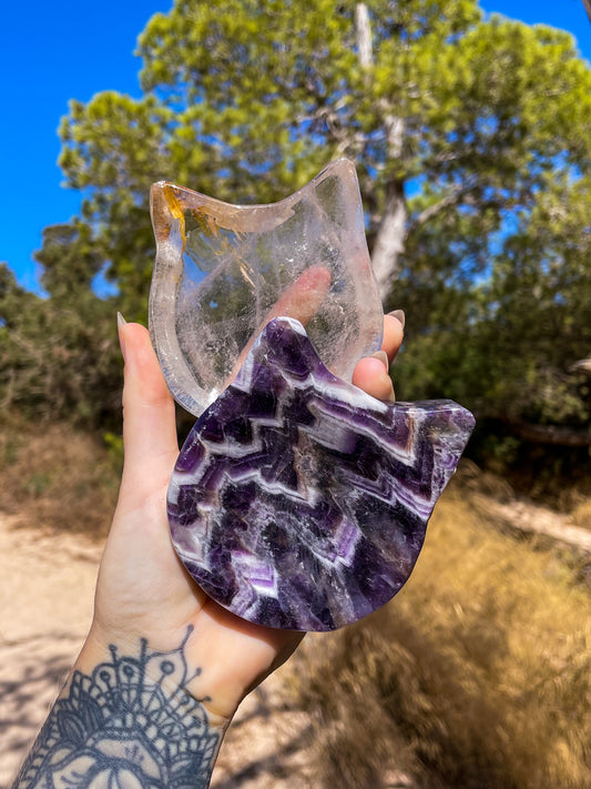A hand holding a cat-shaped crsytal bowls with dreamy amethyst, and clear quartz, with a background of a natural outdoor setting.