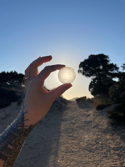 A person's hand holding a clear quartz full moon against a backdrop of trees and a blue sky.