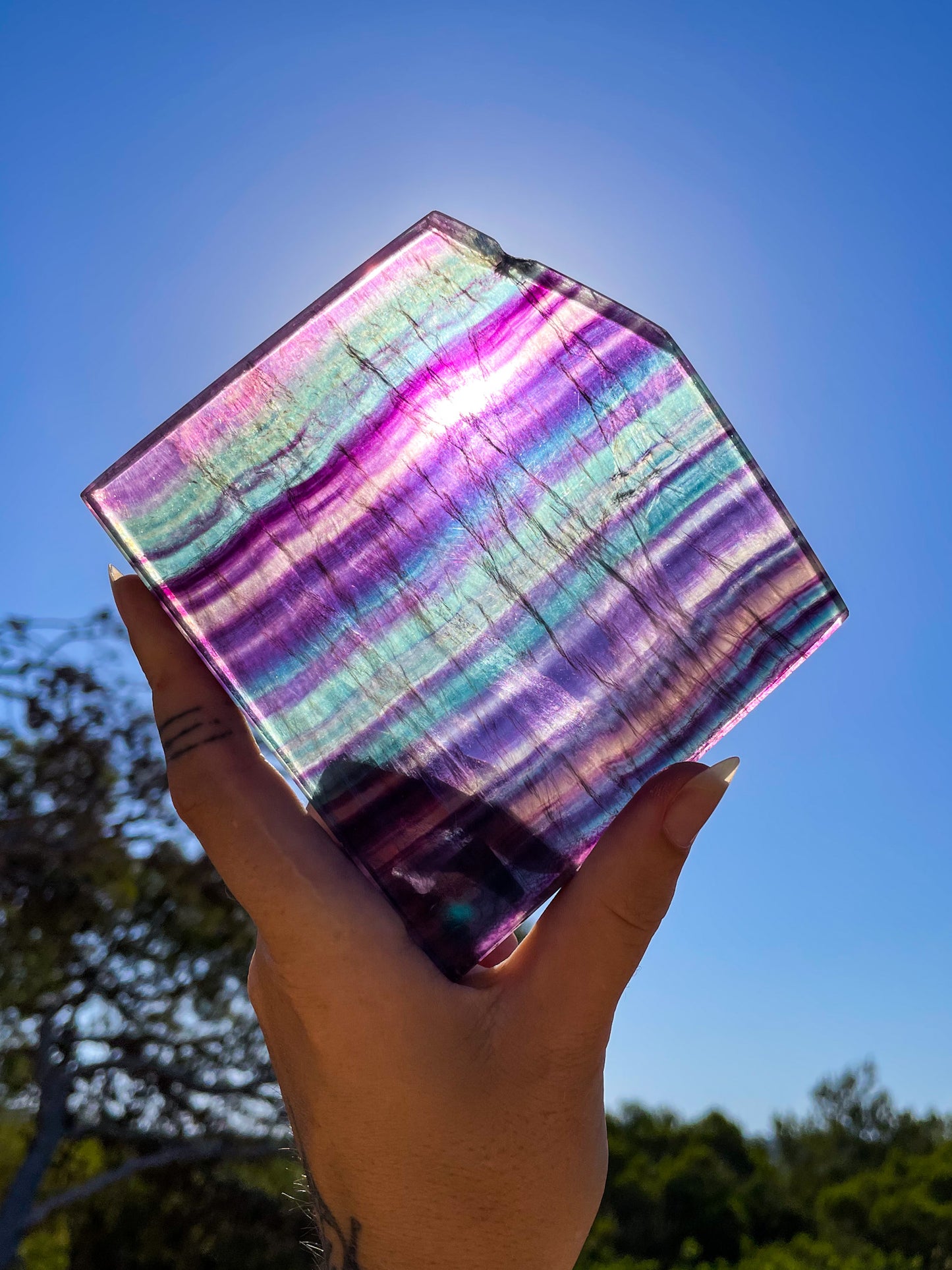 hand holding rainbow fluorite clab up to the sun showing off its rainbow stripes of blues, purples, greens and lilacs